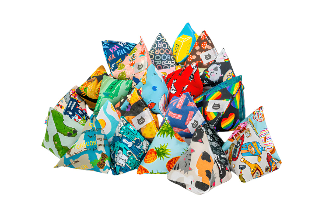 A colorful assortment of pyramid-shaped Happy Sack catnip toys featuring vibrant designs, including food prints, rainbow patterns, and playful animal themes made by Catniptakeout